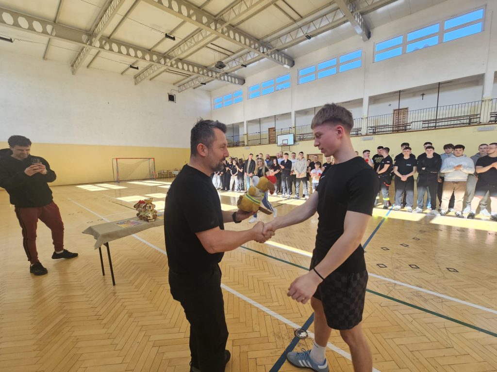 Dwóch mężczyzn w czarnych ubraniach podaje sobie ręce na sali sportowej, jeden z nich trzyma pluszową maskotkę, w tle grupa ludzi stoi wzdłuż ściany.