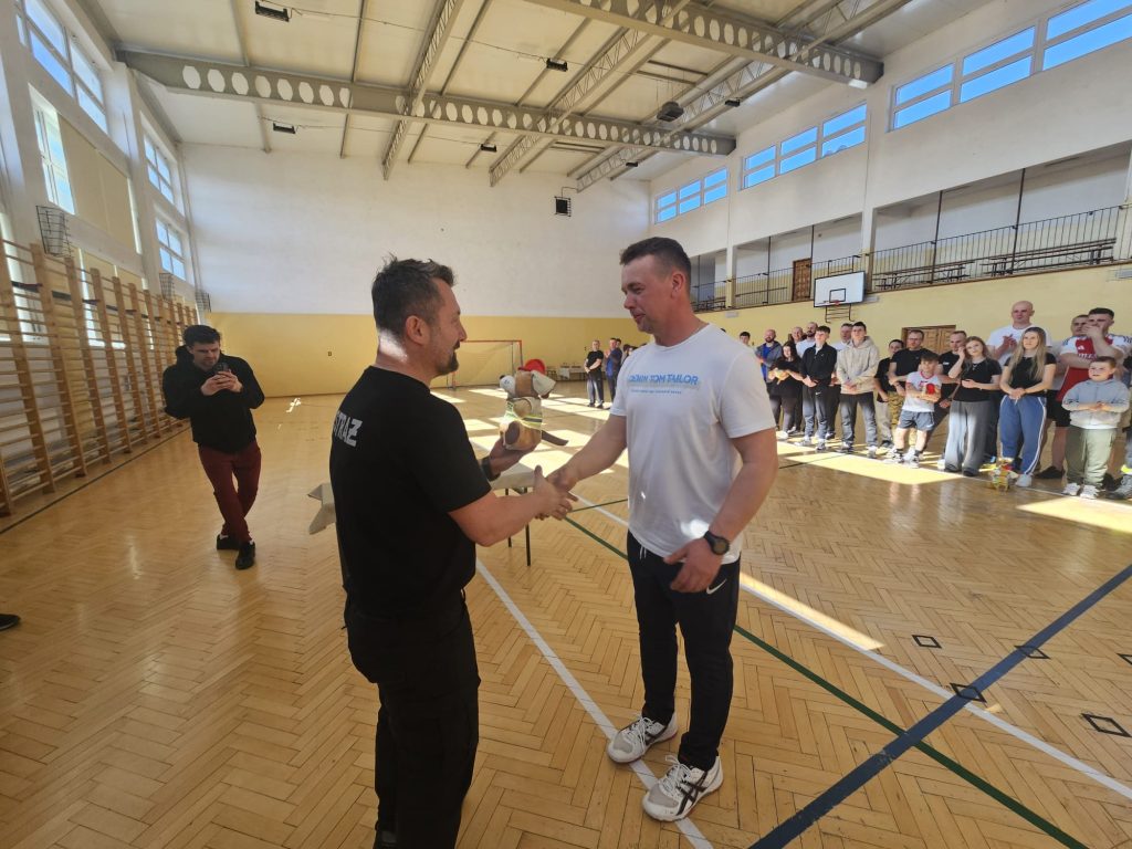 Dwóch mężczyzn podających sobie ręce na parkiecie hali sportowej, jeden trzyma pluszowego psa, w tle grupa osób obserwujących.