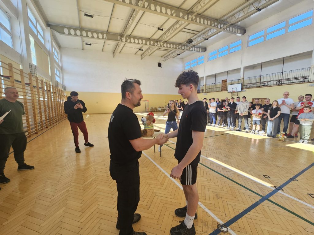 Dwóch mężczyzn ściska dłonie na drewnianej podłodze hali sportowej, w tle stoją ludzie obserwujący wydarzenie przy ścianie z oknami.