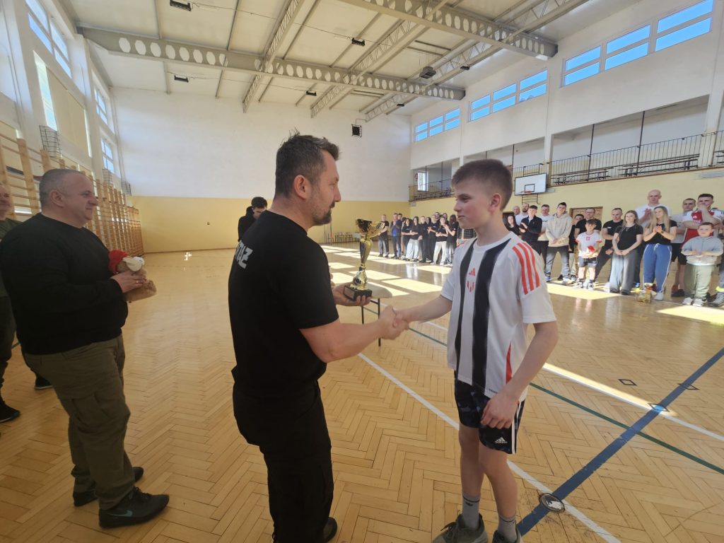 Chłopiec w białej koszulce z czarnymi i czerwonymi pasami odbiera puchar od mężczyzny w czarnej koszulce na tle grupy ludzi w hali sportowej.