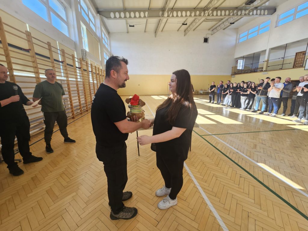 Mężczyzna i kobieta w czarnych ubraniach podają sobie ręce na hali sportowej, mężczyzna trzyma pluszowego psa w czerwonym kapeluszu, w tle stoją ludzie i oklaskują.