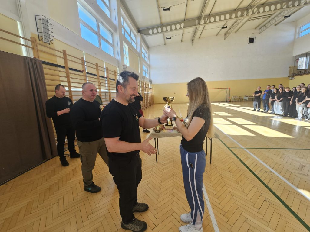 Mężczyzna w czarnej koszulce wręcza puchar kobiecie w niebieskich spodniach dresowych i czarnej koszulce na drewnianej podłodze hali sportowej, w tle ludzie stojący przy ścianie.