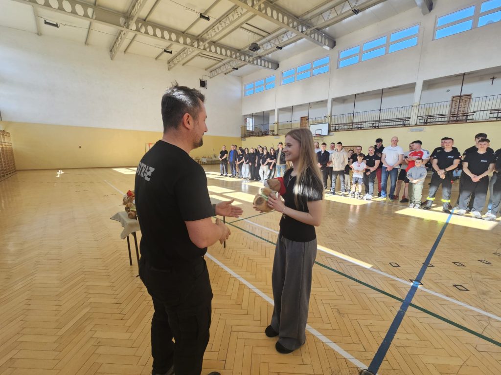 Mężczyzna w czarnej koszulce z napisem "STRAŻ" wręcza dziewczynie pluszowego misia na jasnej drewnianej podłodze hali sportowej, w tle stoi grupa ludzi.