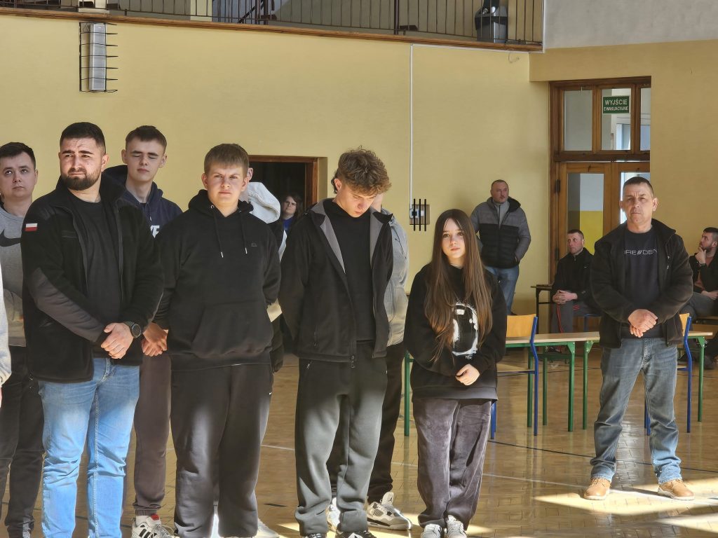 Grupa osób w sportowych ubraniach stoi w hali z żółtymi ścianami i drewnianą podłogą, w tle stoły i drzwi z zielonym napisem "WYJŚCIE EWAKUACYJNE".