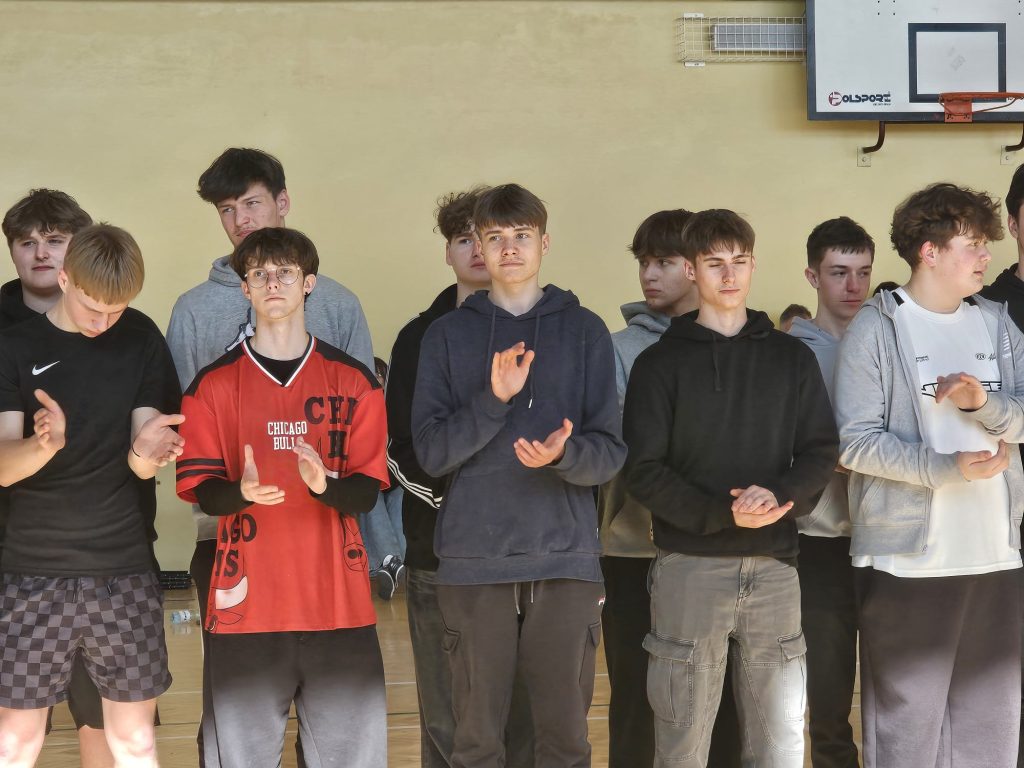 Grupa nastoletnich chłopców ubranych w sportowe ubrania stoi na hali sportowej, niektórzy klaszczą, w tle jasna ściana i tablica do koszykówki.