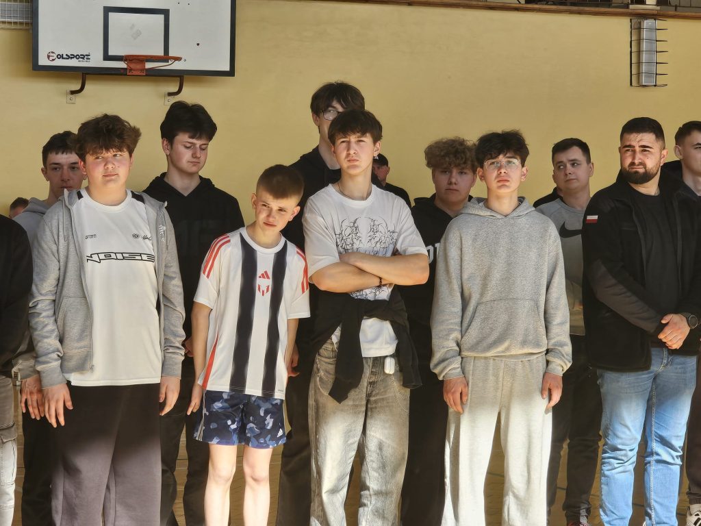 Grupa młodych mężczyzn stoi w hali sportowej pod tablicą do koszykówki, ubrani w sportowe i casualowe ubrania, na żółtej ścianie widoczna lampa.