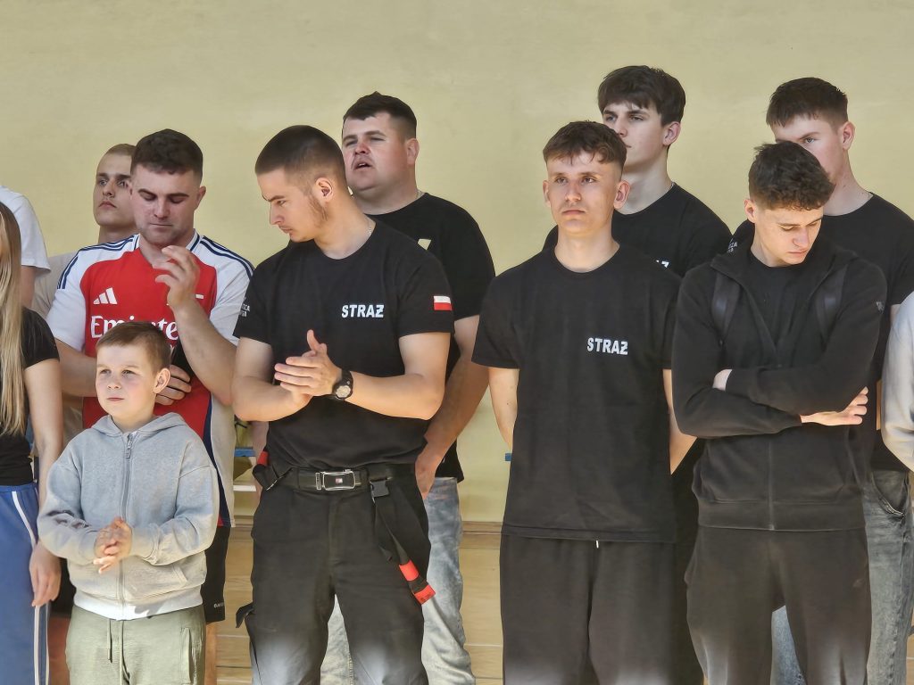 Grupa młodych mężczyzn i chłopiec, większość w czarnych ubraniach z napisem "STRAŻ", stojący na jasnym tle w pomieszczeniu.