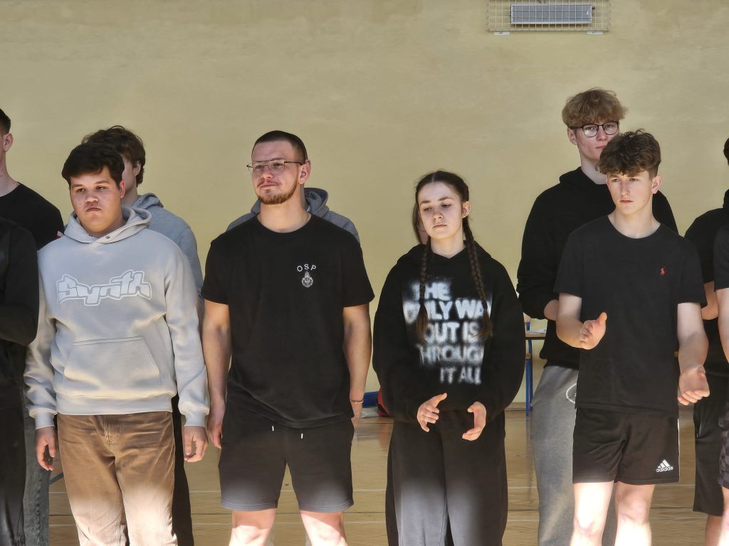 Grupa młodych osób stojących na tle żółtej ściany, ubranych w czarne i jasne sportowe ubrania, niektórzy mają okulary i warkocze.