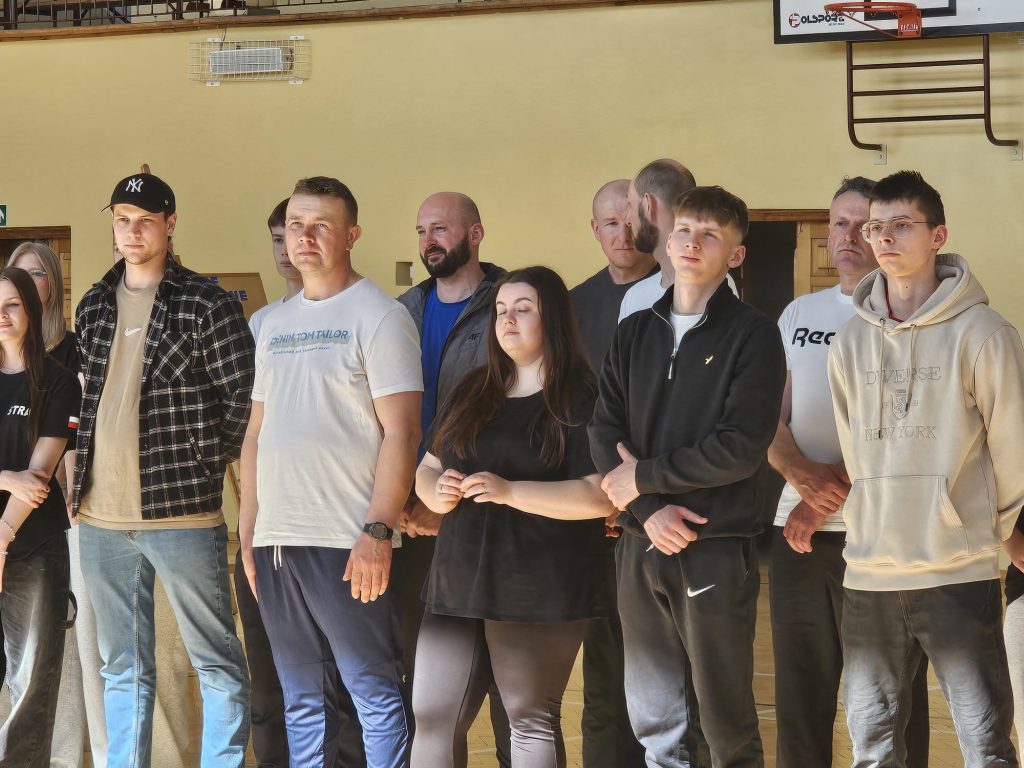 Grupa ludzi w sportowej odzieży stojąca w hali sportowej, na ścianie żółta farba, w tle kosz do koszykówki i drabinka gimnastyczna.