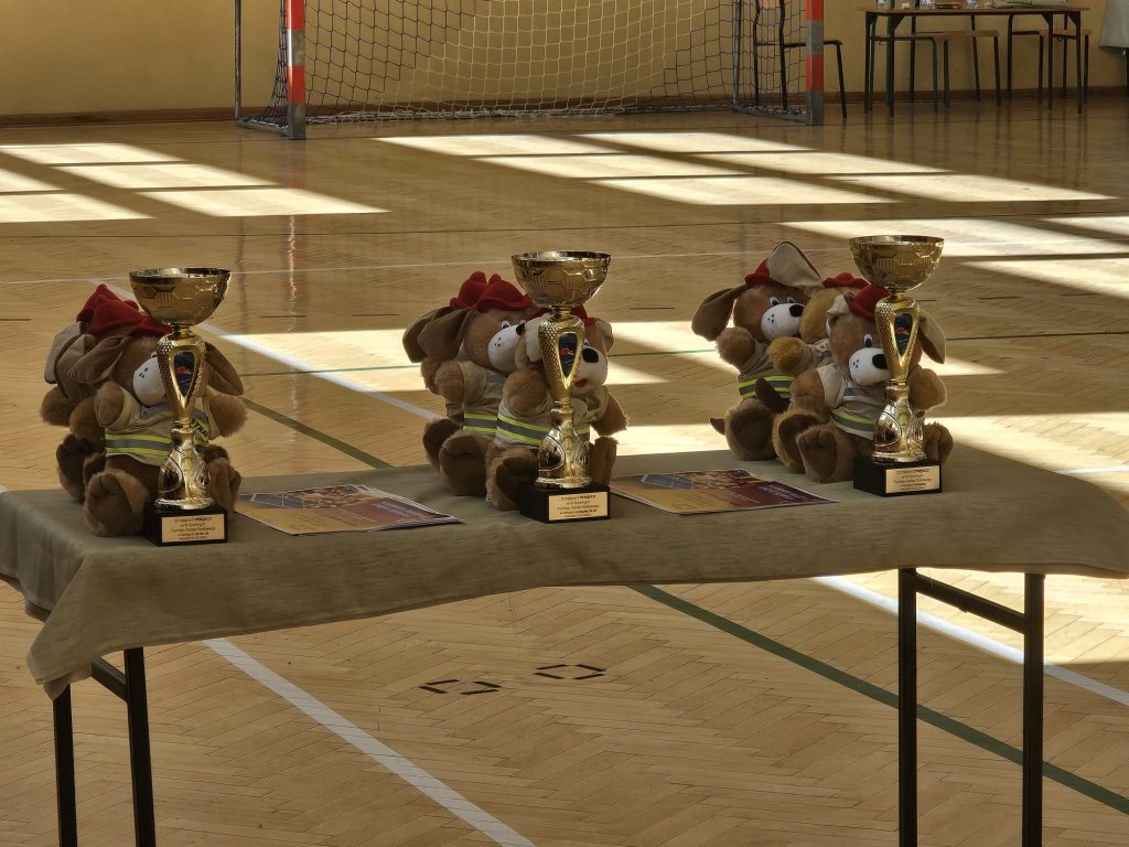 Trzy złote puchary i pluszowe misie w ubraniach strażackich na stole w sali sportowej z drewnianą podłogą i bramką.