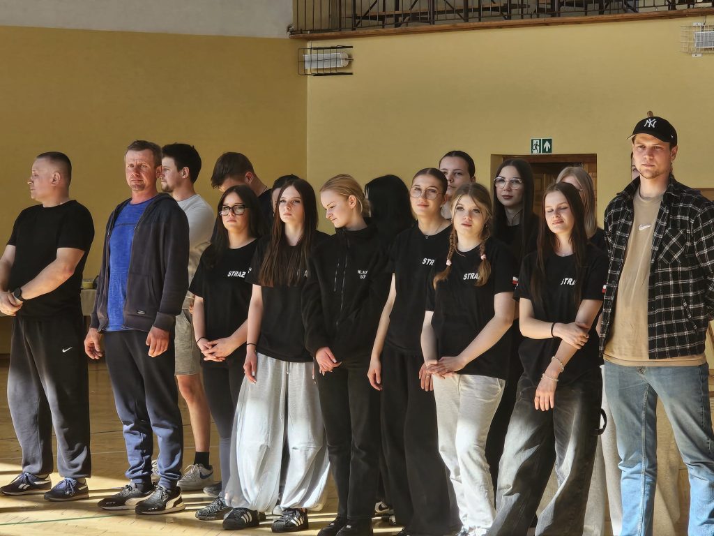 Grupa ludzi stoi w rzędzie na sali gimnastycznej, większość w czarnych ubraniach z napisem "STRAŻ", na żółtej ścianie drzwi i znak ewakuacyjny.