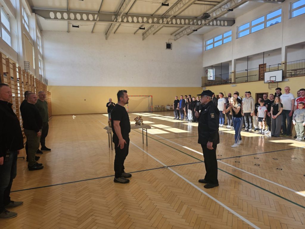 Ludzie ustawieni w dwóch rzędach w hali sportowej, dwóch mężczyzn stoi naprzeciwko siebie przy stole z pucharami, na ścianie okna i bramka do piłki ręcznej.