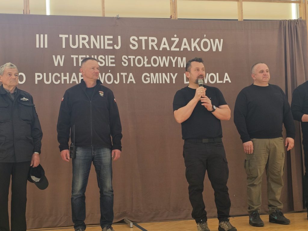 Czterech mężczyzn w ciemnych ubraniach stoi na parkiecie przed brązową kurtyną z białym napisem. Jeden z nich trzyma mikrofon.