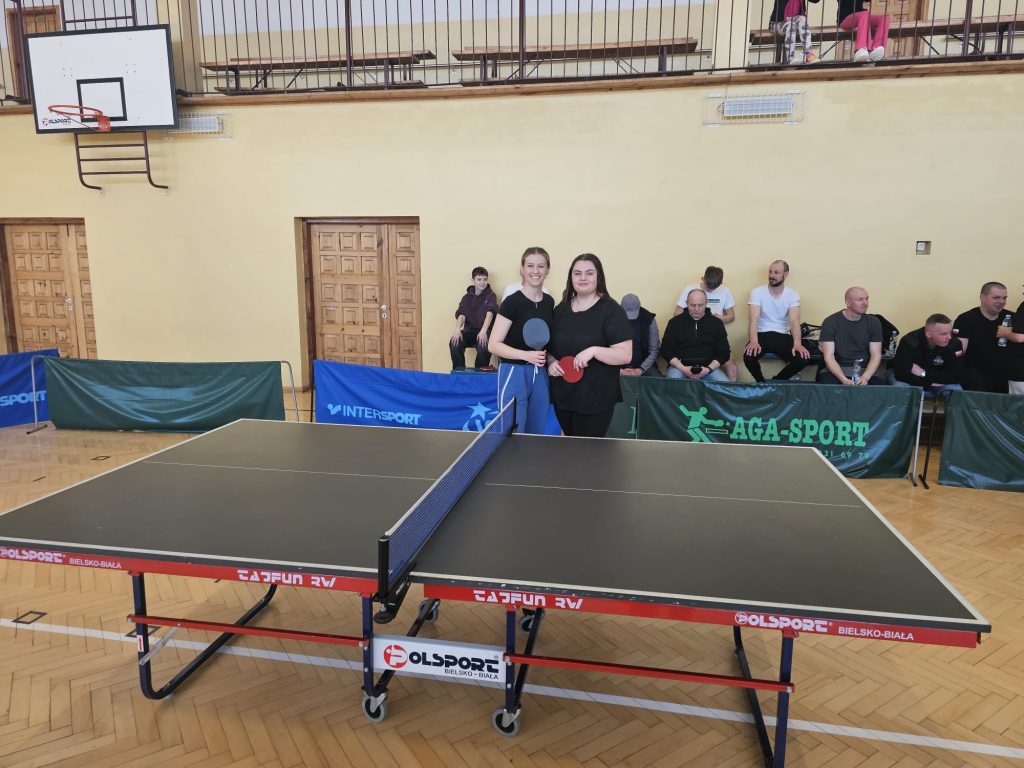 Stół do tenisa stołowego na hali sportowej, dwie kobiety trzymające rakietki stoją za stołem, w tle siedzą ludzie na ławce przy ścianie z drzwiami.