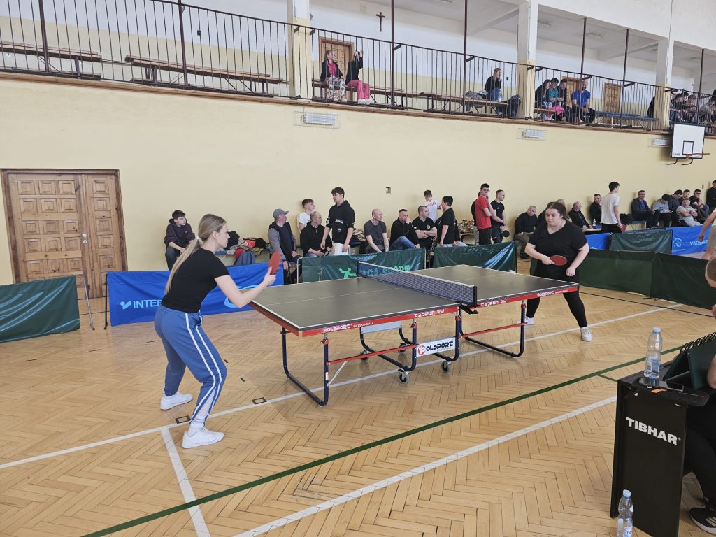 Dwie kobiety grające w tenisa stołowego, drewniana podłoga hali sportowej, widownia z siedzącymi ludźmi, zielone i niebieskie bandy ochronne.