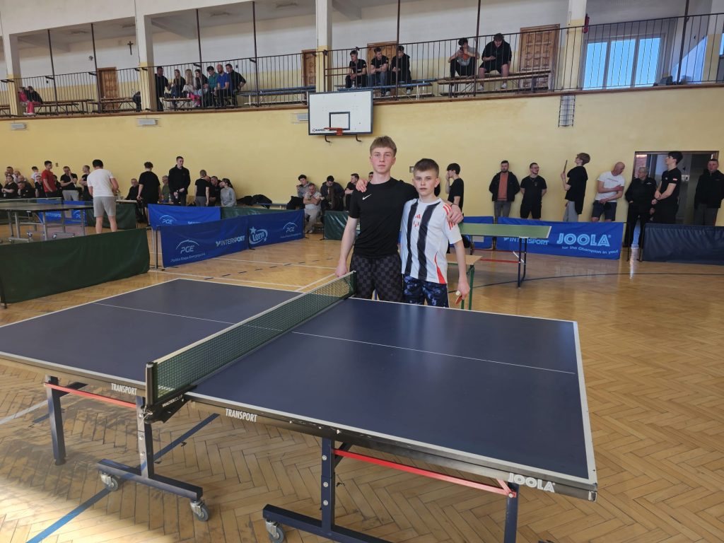 Dwoje chłopców w sportowych strojach stoi przy stole do tenisa stołowego na hali sportowej, w tle widoczni kibice i inni zawodnicy.