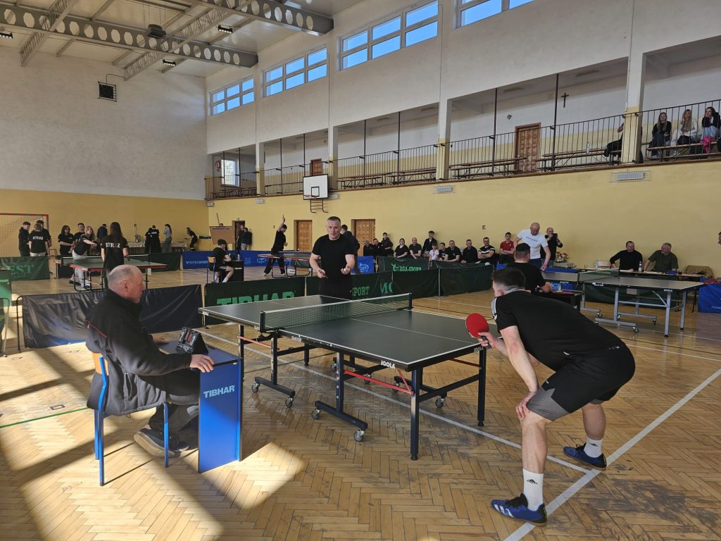 Hala sportowa z kilkoma stołami do tenisa stołowego, zawodnicy w czarnych strojach, sędzia przy stołach z licznikiem, drewniana podłoga, widownia z tyłu.