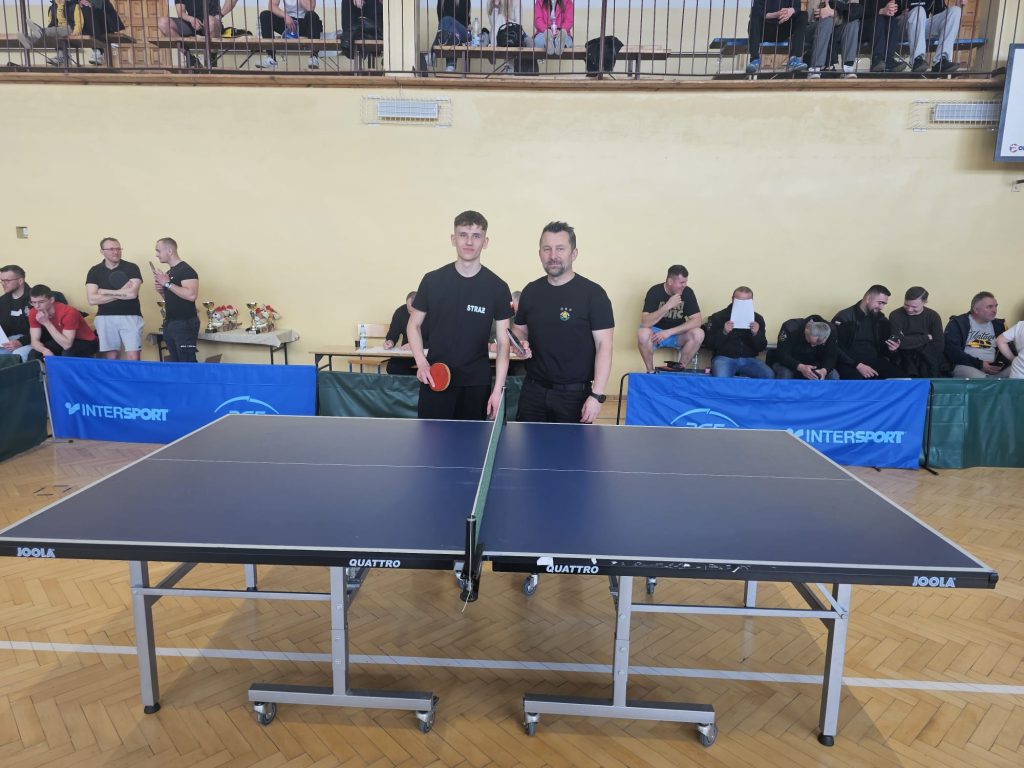 Dwaj mężczyźni w czarnych koszulkach stoją za niebieskim stołem do tenisa stołowego na drewnianej podłodze, w tle widoczna grupa osób siedzących i stojących.