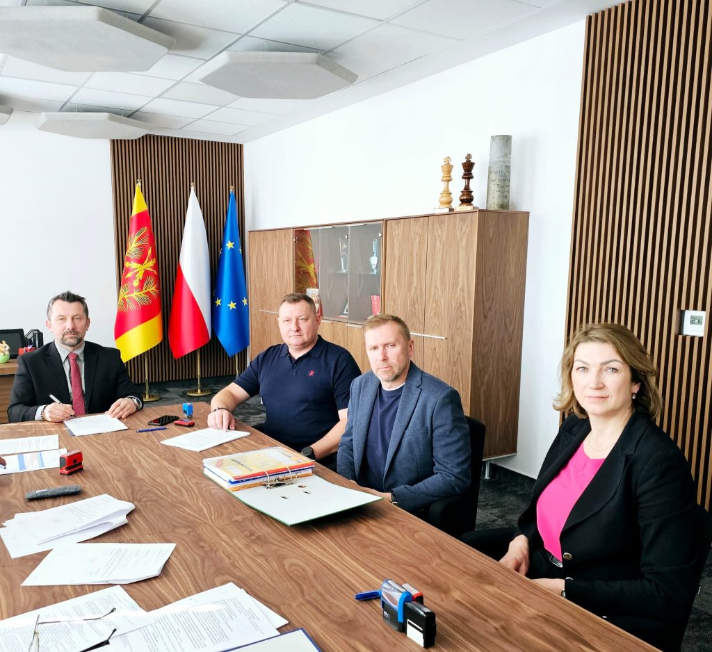 Cztery osoby siedzące przy drewnianym stole konferencyjnym, w tle trzy flagi i szafka z trofeami, na szafce drewniane figury szachowe i cylindryczny przedmiot.