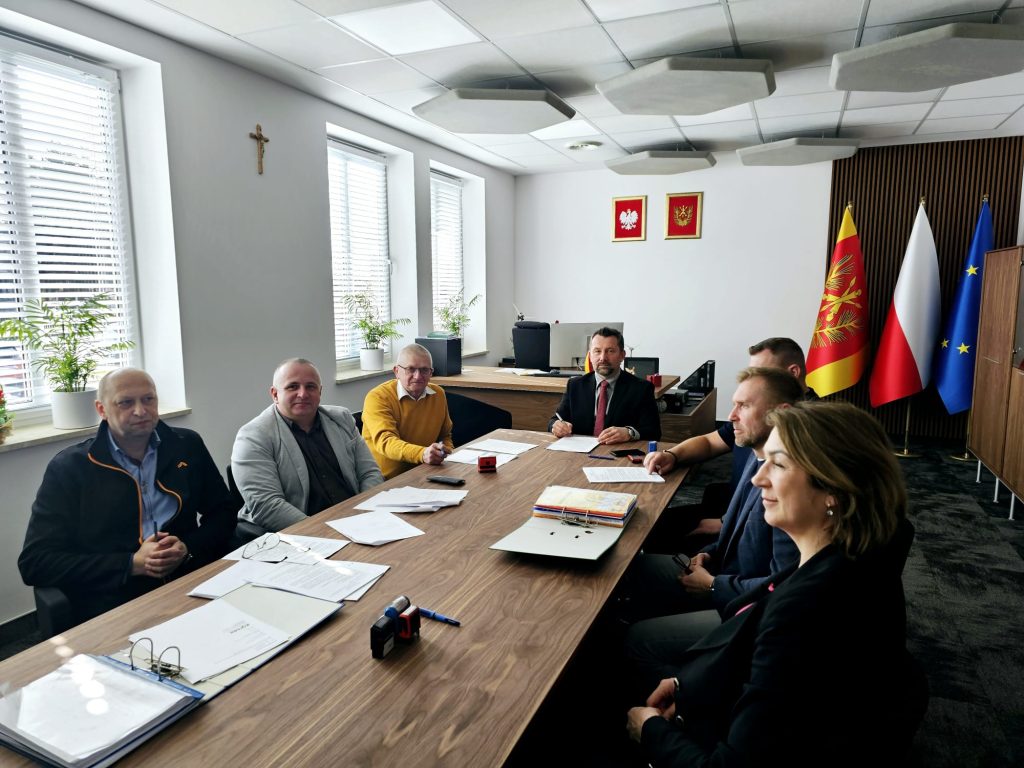 Siedem osób przy drewnianym stole w jasnym biurze z trzema oknami, na ścianie krzyż, godła Polski i województwa, trzy flagi: województwa, Polski i Unii Europejskiej.