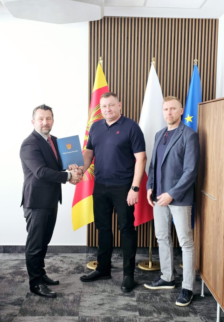 Trzech mężczyzn na tle trzech flag, dwóch z nich podaje sobie ręce, jeden trzyma niebieską teczkę z napisem "Wójt Gminy Dzwola".