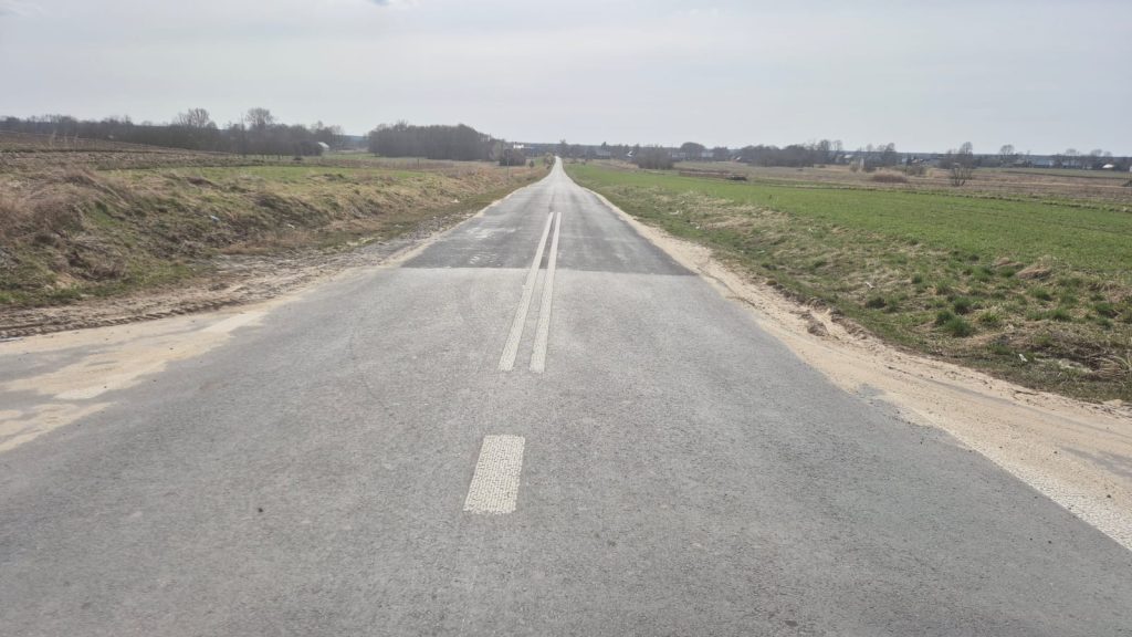 Asfaltowa droga z podwójną linią ciągłą na środku, po obu stronach pola i łąki, w tle drzewa i zabudowania pod szarym niebem.