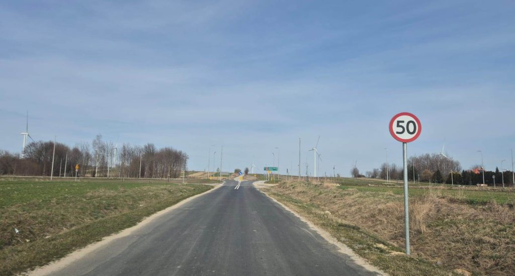 Asfaltowa droga z znakiem ograniczenia prędkości 50 km/h po prawej stronie, po bokach trawa i pola, w tle wiatraki i drzewa pod niebieskim niebem.