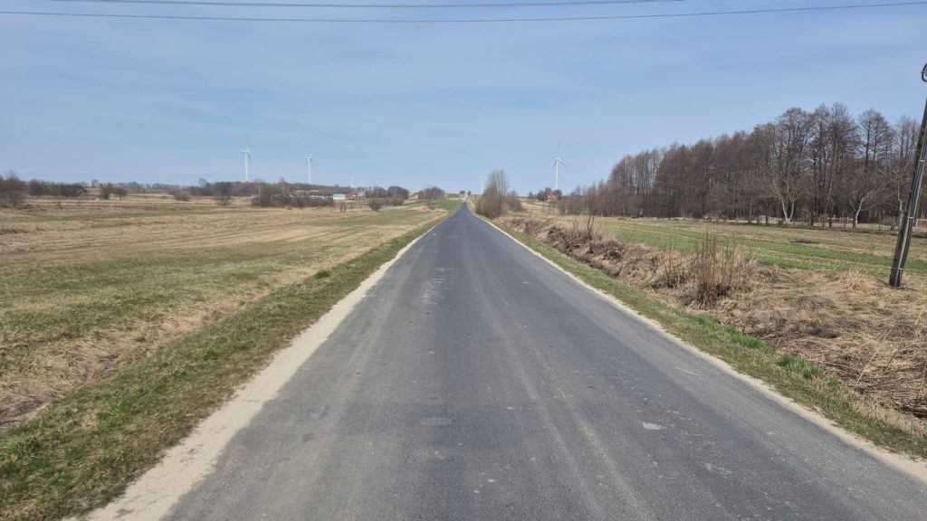 Asfaltowa droga biegnąca przez pola z suchą trawą, po prawej stronie las z drzewami bez liści, na horyzoncie wiatraki i jasne niebo.