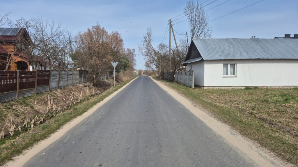 Wąska asfaltowa droga z poboczami z piasku, po lewej drewniany płot i drzewa bez liści, po prawej biały dom z metalowym dachem i trawnik. W tle bezlistne drzewa i niebieskie niebo.