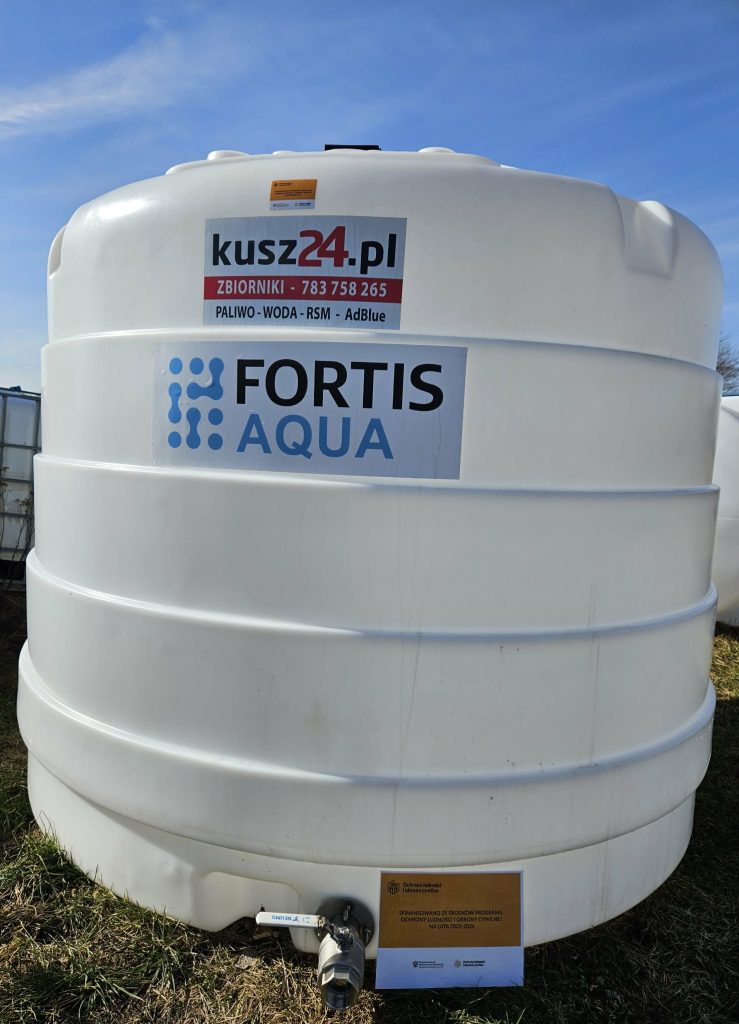 Biały plastikowy zbiornik z naklejkami "kusz24.pl", "FORTIS AQUA" oraz informacją o finansowaniu programu ochrony ludności i obrony cywilnej, zawór u dołu, trawa i niebieskie niebo.