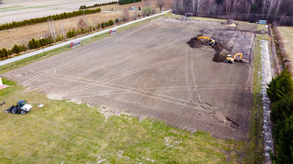 duży plac ziemi z widocznymi śladami kół, dwie koparki pracujące na prawo, traktor na trawie po lewej, w tle drzewa i droga z przystankami autobusowymi
