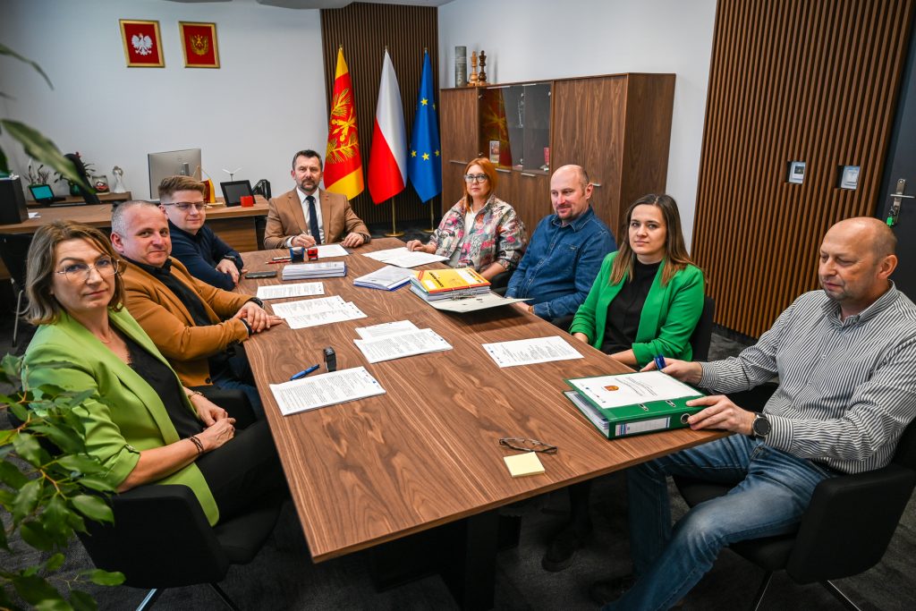 Osiem osób przy drewnianym stole konferencyjnym, dokumenty i segregatory, w tle trzy flagi i dwa czerwone godła na białej ścianie.