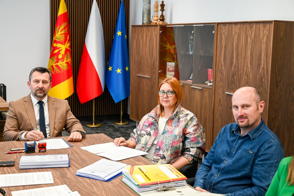 Trzy osoby siedzące przy drewnianym stole z dokumentami, w tle trzy flagi: żółto-czerwona, biało-czerwona i niebieska z gwiazdami, za nimi drewniana szafa z przeszklonymi drzwiami.