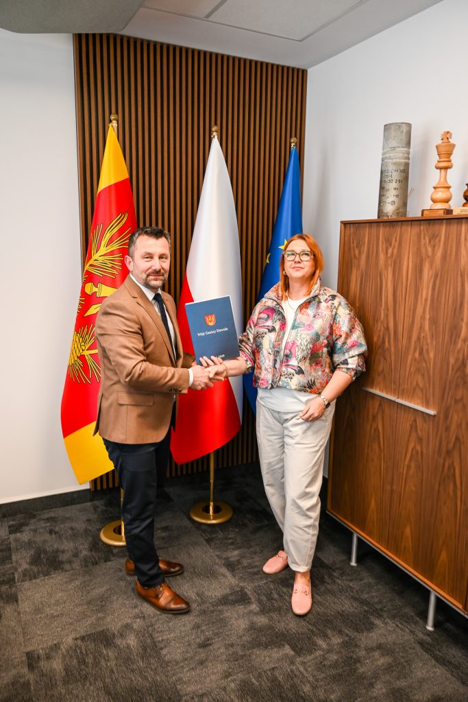 Mężczyzna w brązowej marynarce i kobieta w kolorowej kurtce ściskają sobie dłonie, obok trzy flagi i drewniana szafka z dekoracjami.