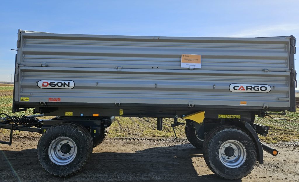 Przyczepa rolnicza szara z metalowymi burtami, duże czarne opony z szarymi felgami, oznaczenia D60N i CARGO, żółte i czerwone naklejki ostrzegawcze, tło pola i niebieskie niebo.