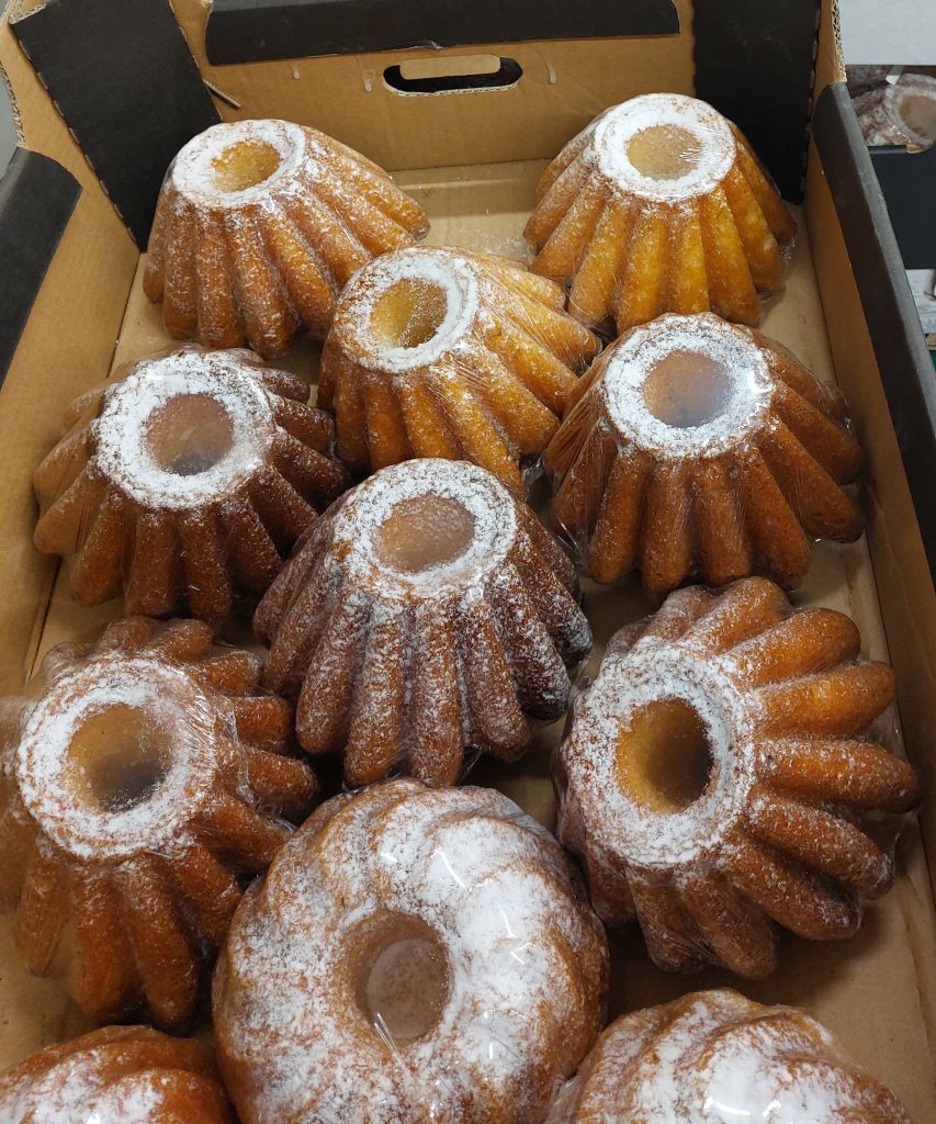 Pudełko z kilkunastoma babkami o kształcie stożkowym pokrytymi cukrem pudrem, zapakowanymi w przezroczystą folię. Babki mają złocisto-brązowy kolor.