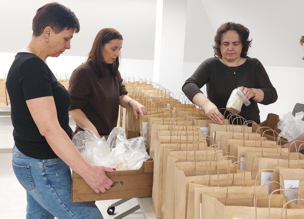 Trzy kobiety pakujące plastikowe woreczki z białą zawartością do papierowych toreb stojących na stołach w jasnym pomieszczeniu.