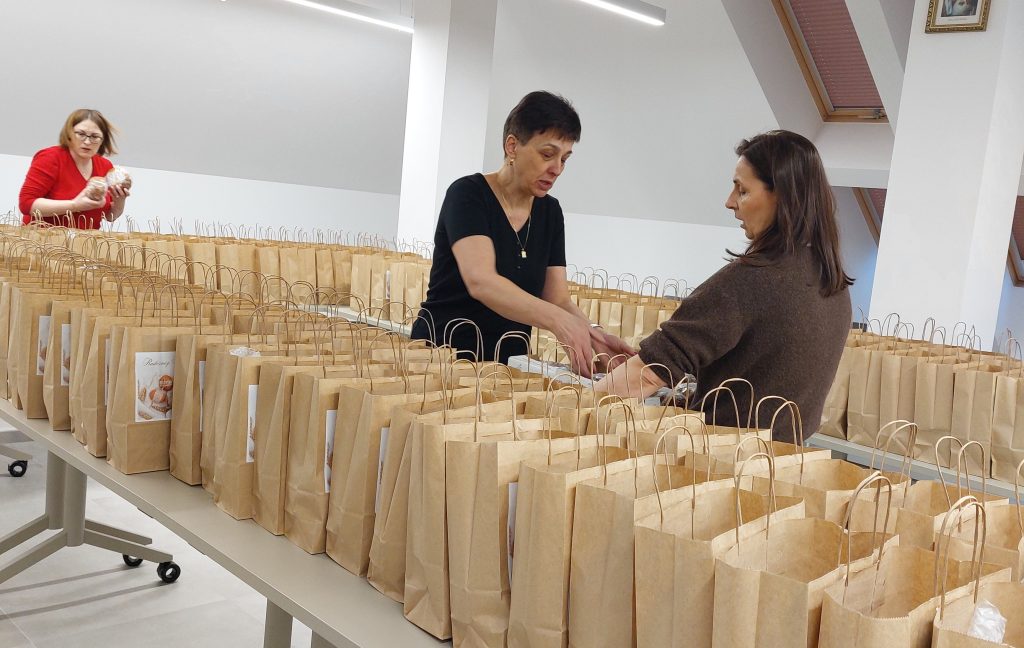 Trzy kobiety przy stołach z dużą ilością brązowych papierowych torebek z uchwytami, jasne wnętrze z oknami dachowymi i obrazem na ścianie.