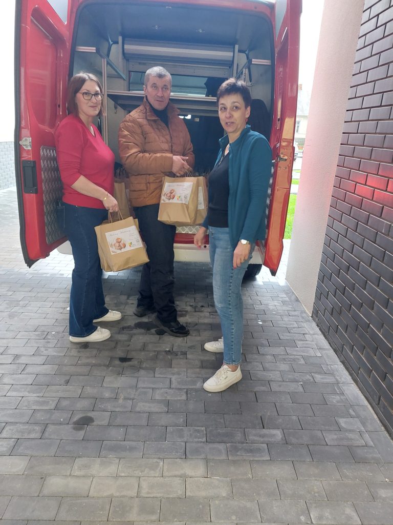 Trzy osoby stoją przy otwartych drzwiach czerwonego pojazdu dostawczego, trzymające brązowe torby papierowe z białymi etykietami, na szarej kostce brukowej.