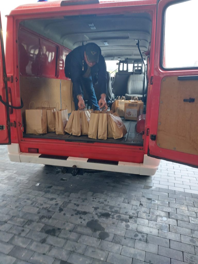 Czerwony van z otwartymi tylnymi drzwiami, w środku mężczyzna układający brązowe papierowe torby na podłodze pojazdu.