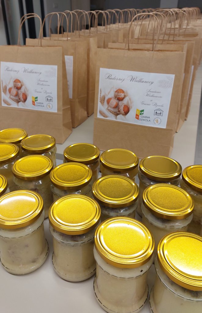 Słoiki z jasnym kremowym wnętrzem i złotymi nakrętkami na białym stole, za nimi brązowe torby papierowe z białymi etykietami ozdobionymi obrazkiem jajek i tekstem.