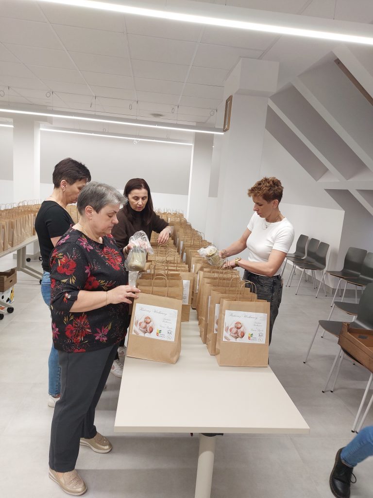 Cztery kobiety stoją przy dużym białym stole, pakują małe torby papierowe z uchwytami i naklejkami z napisami i zdjęciami ciastek, w jasnym pomieszczeniu z krzesłami.