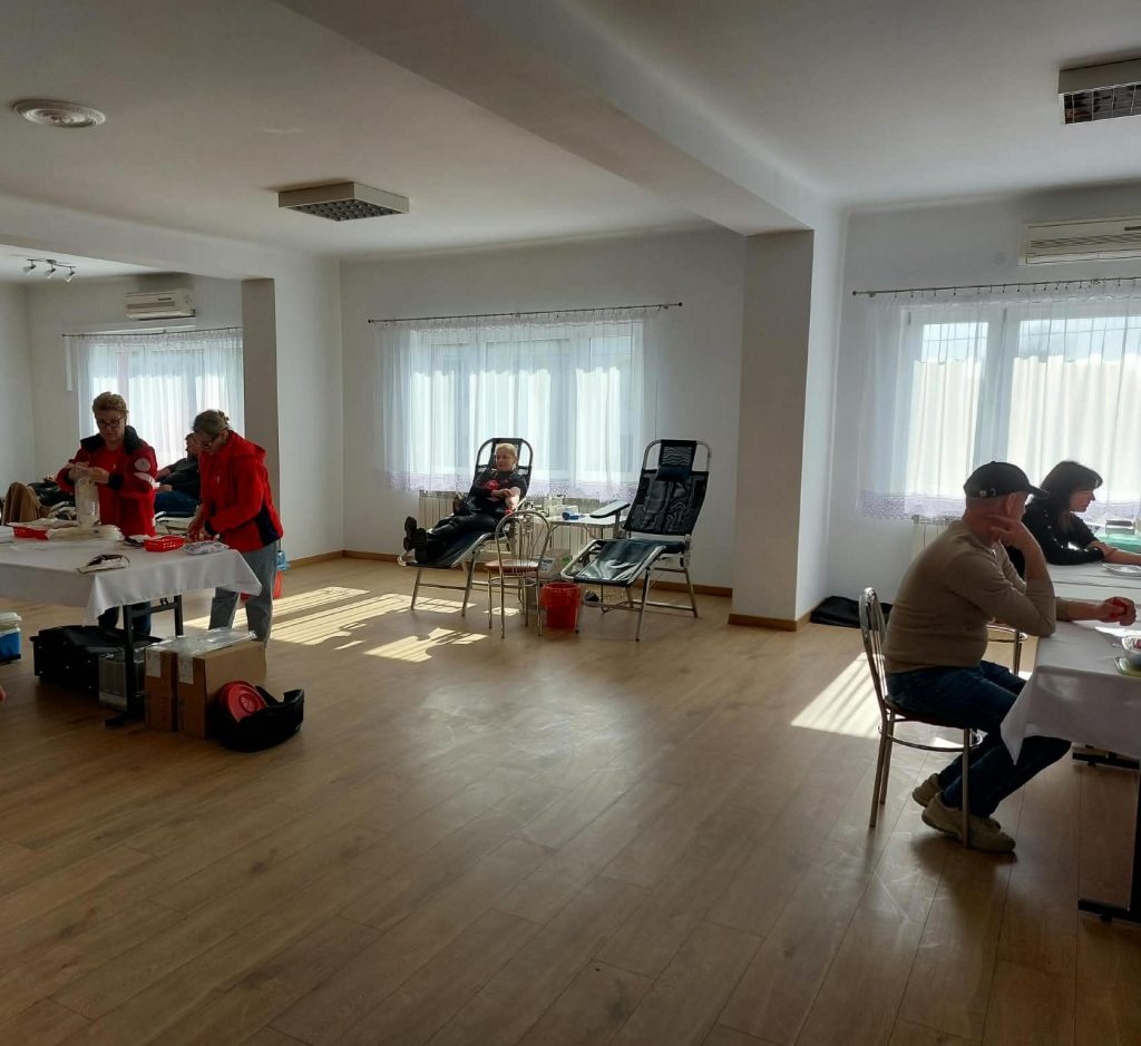 Jasne pomieszczenie z drewnianą podłogą, dwie osoby w czerwonych kurtkach przy stole z białym obrusem, osoba leżąca na fotelu, mężczyzna i kobieta siedzący przy innym stole.