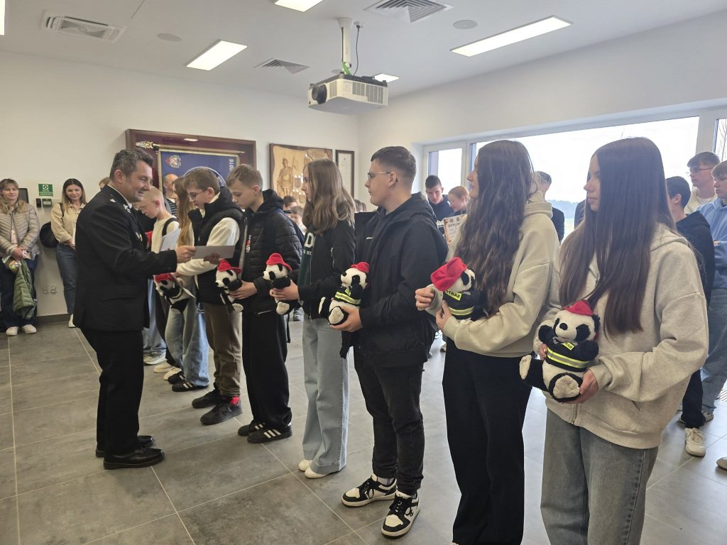 Grupa nastolatków stoi w rzędzie w jasnym pomieszczeniu, każdy trzyma pluszowego misia w czerwonej czapce i kamizelce, mężczyzna w mundurze wręcza im dokumenty.