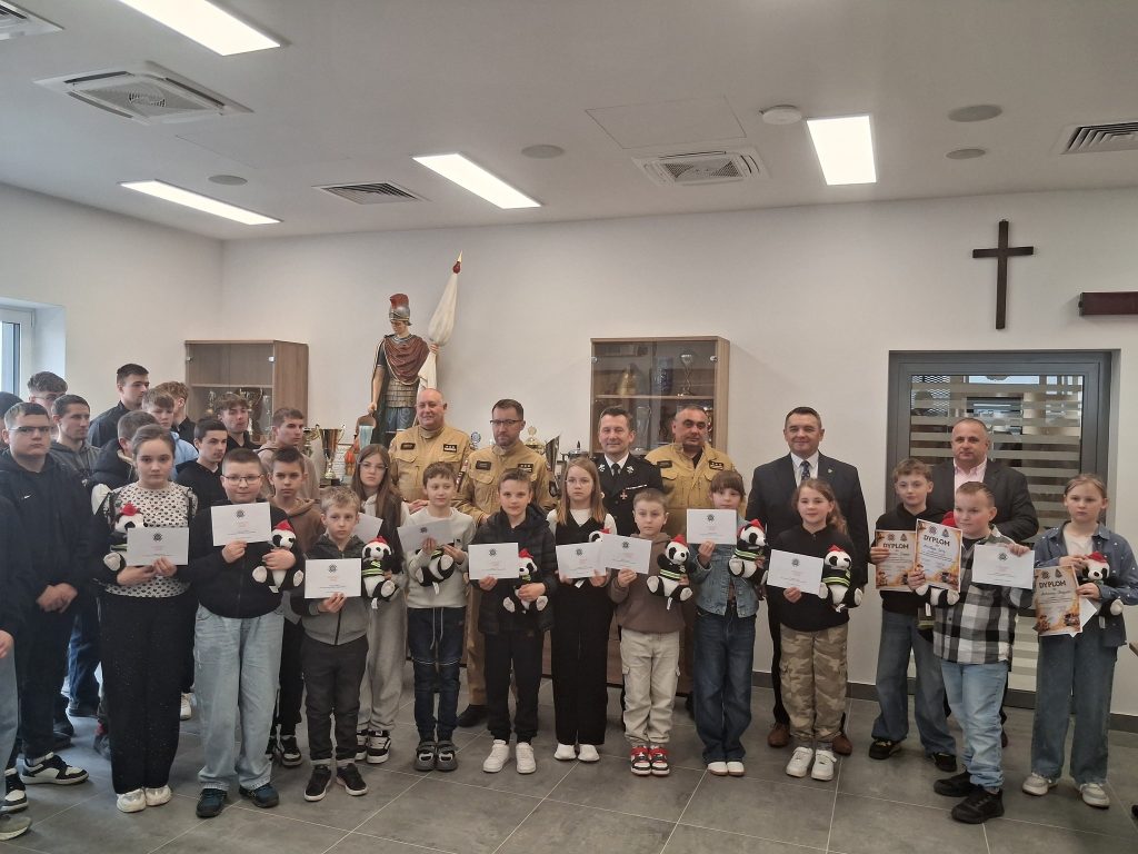 Grupa dzieci i dorosłych na tle białej ściany z krzyżem, dzieci trzymają dyplomy i pluszowe pandy, w tle statua wojownika i szafki z trofeami.