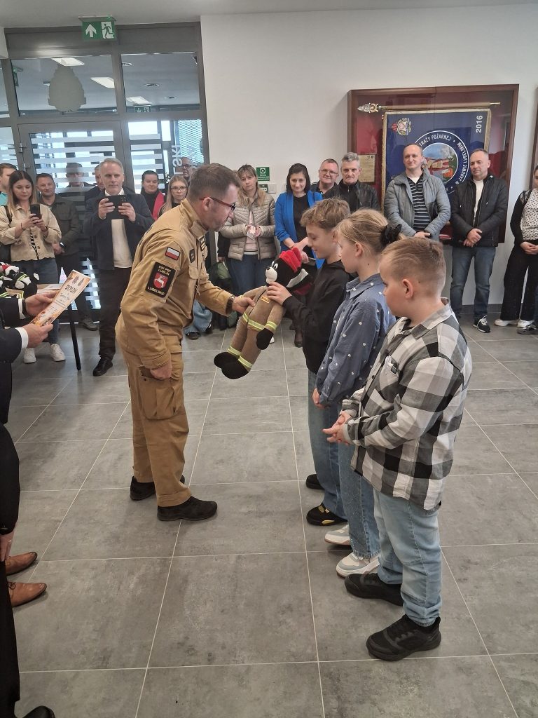 Mężczyzna w beżowym mundurze przekazuje pluszaka w stroju strażaka chłopcu, obok stoją dwójka dzieci, w tle grupa osób obserwujących wydarzenie.