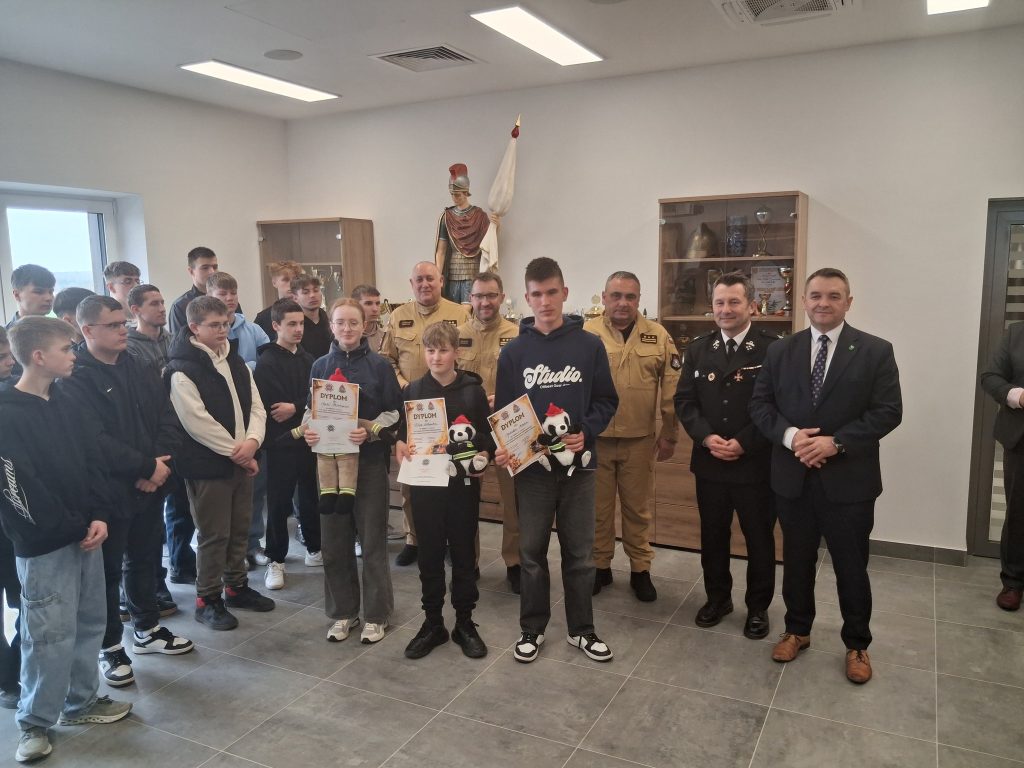 Grupa osób w pomieszczeniu, w tym młodzież trzymająca dyplomy i pluszaki, oraz dorośli w mundurach i garniturach, na tle szafek z trofeami i figurą żołnierza.