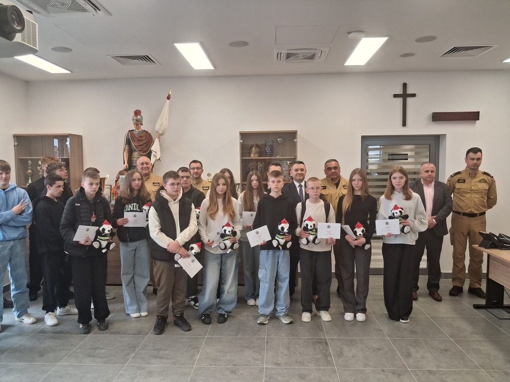 Grupa młodzieży i dorosłych w jasnym pomieszczeniu, część osób trzyma białe koperty i pluszowe pandy w czapkach czerwonych i żółtych kamizelkach, w tle drewniana szafa i krzyż.