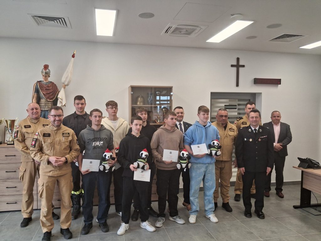 Grupa mężczyzn w mundurach i ubraniach cywilnych w jasnym pomieszczeniu, część trzyma pluszowe misie i dyplomy, na ścianie drewniany krzyż i szafka z trofeami.