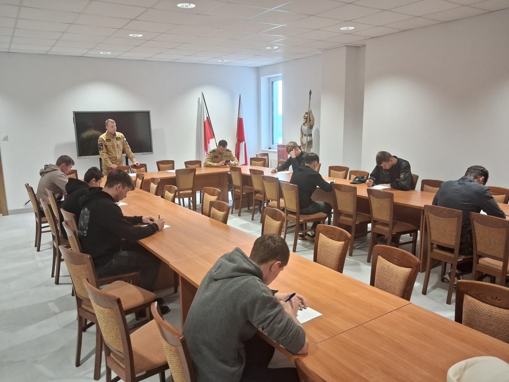 Mężczyźni siedzący przy drewnianych stołach, piszący na kartkach, w jasnym pomieszczeniu z białymi ścianami, flagami i figurą rycerza przy oknie.